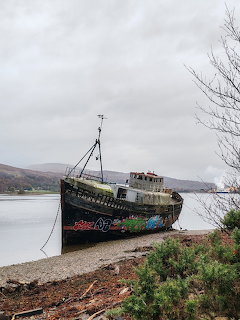 Graffiti-covered shipwreck beached on a rocky shore with hills and overcast sky in Scotland