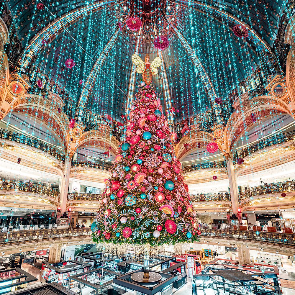 Tall Christmas tree with red ornaments in multi-story shopping mall interior under blue light canopy