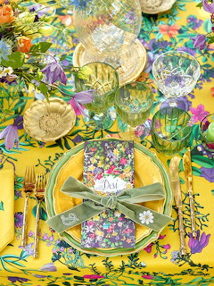 Overhead view of a floral table setting with yellow tablecloth, patterned napkin, gold cutlery and colorful glasses