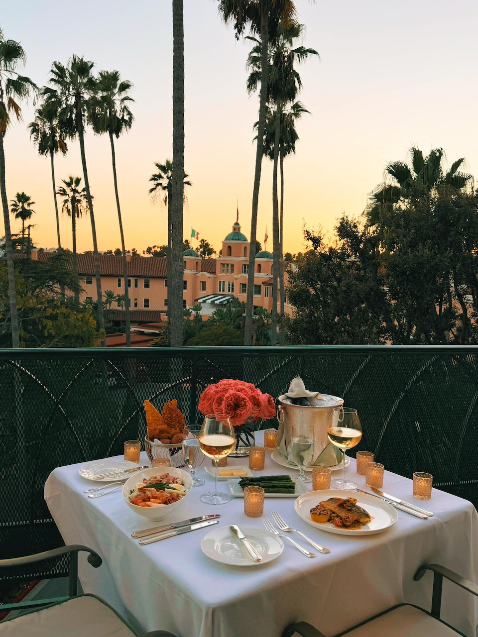 Breakfast table on balcony at sunset with palm trees and historic building silhouettes in warm golden light