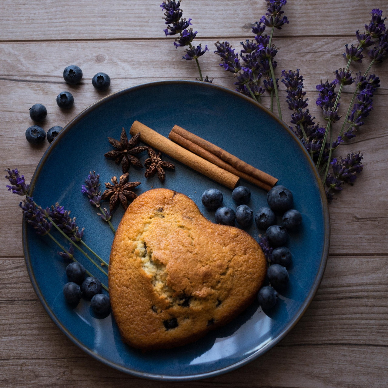Heart-shaped blueberry cake garnished with fresh blueberries, cinnamon, and lavender on a blue plate