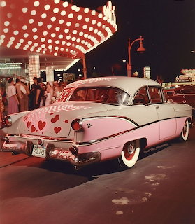 Pink and white 1950s vintage car decorated with red hearts parked under illuminated canopy at night