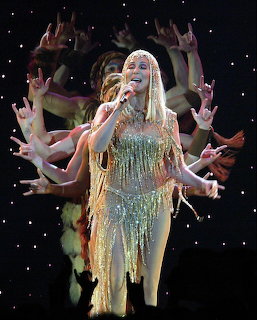 Cher singing on stage in a glittering fringed costume with dancers' hands reaching out behind her