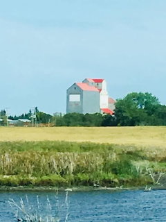 Grain elevator with orange roof behind golden wheat field and lake in rural Saskatchewan