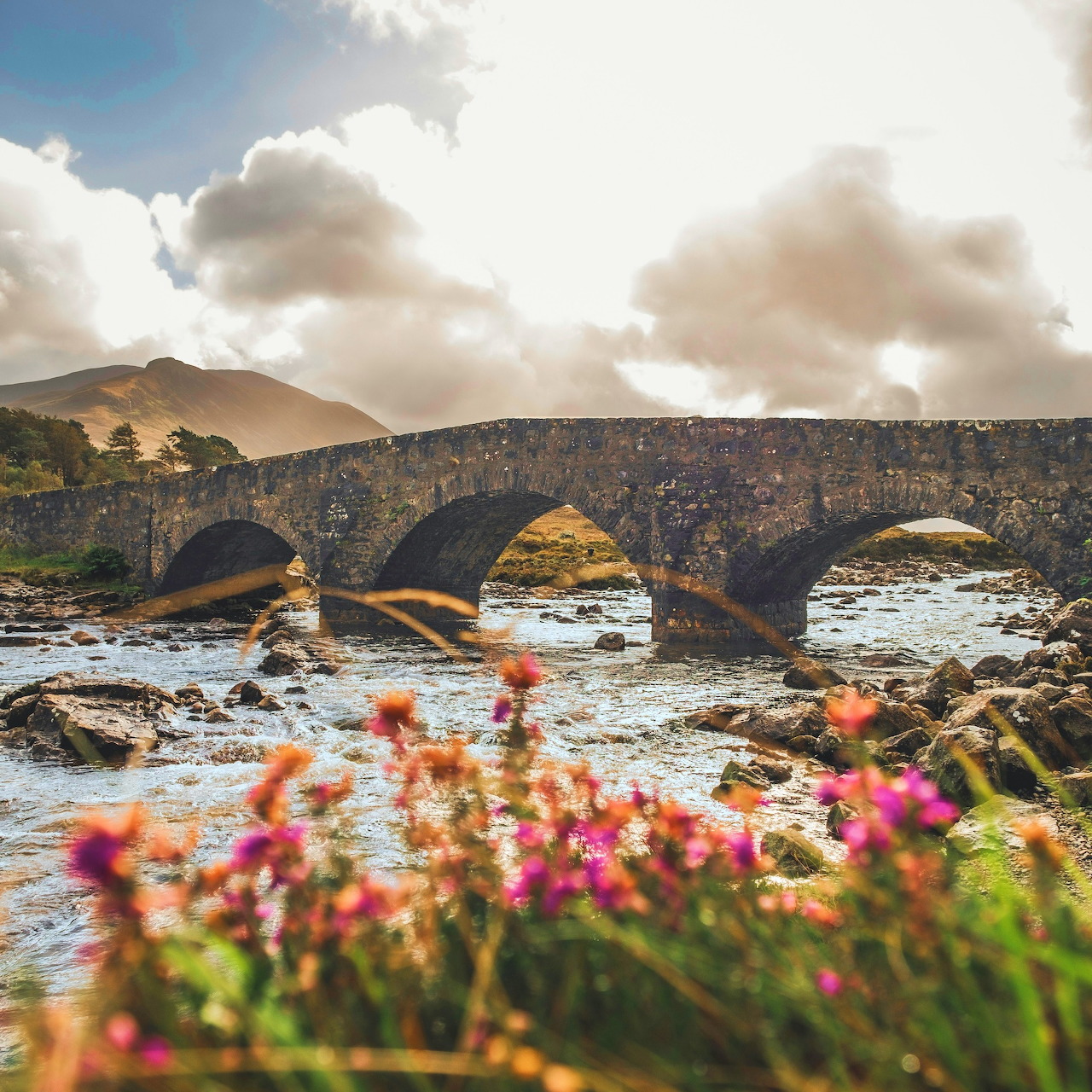 Old stone arch bridge over a rocky river with mountains and clouds on the Isle of Skye, Scotland