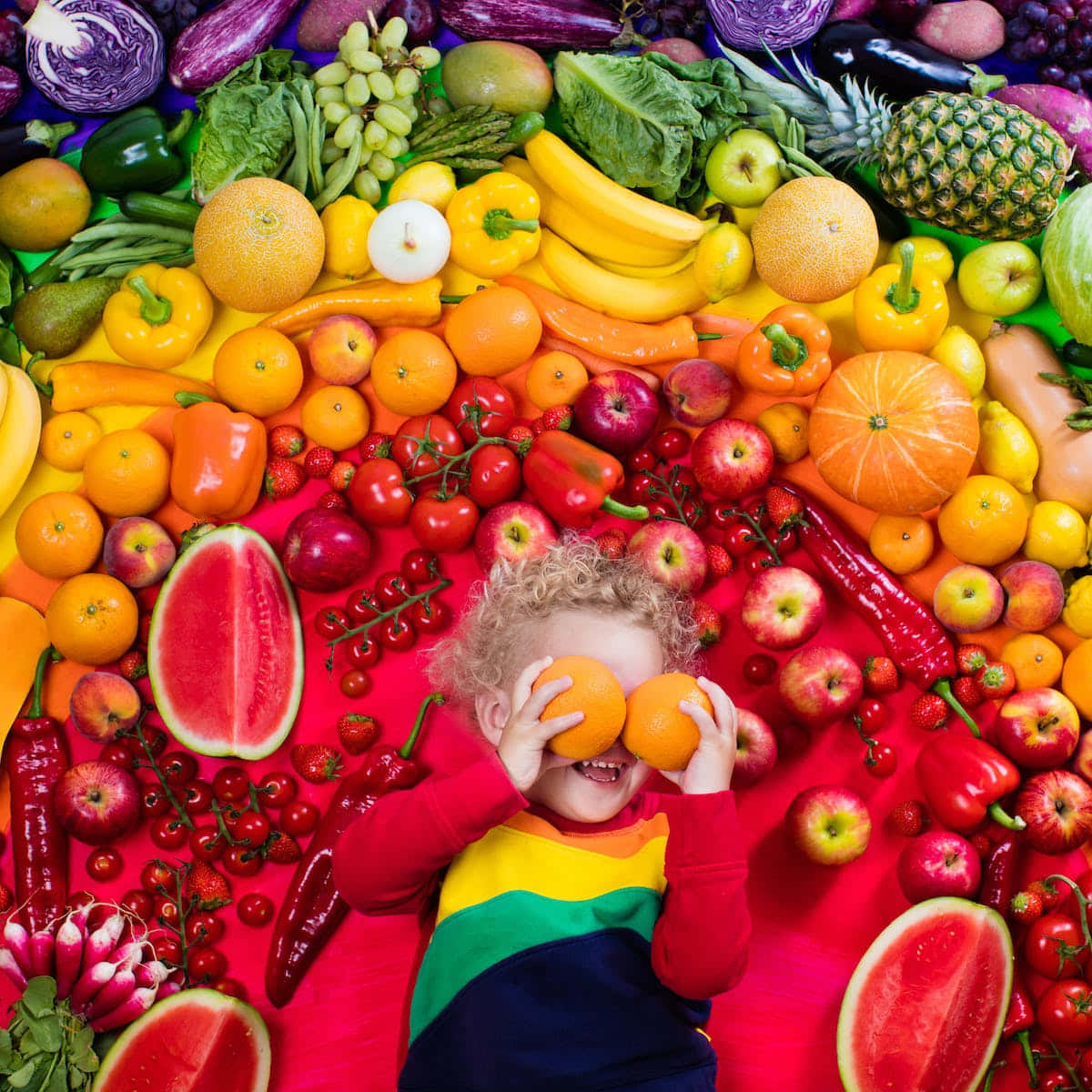 Child surrounded by rainbow-arranged fruits and vegetables holding oranges