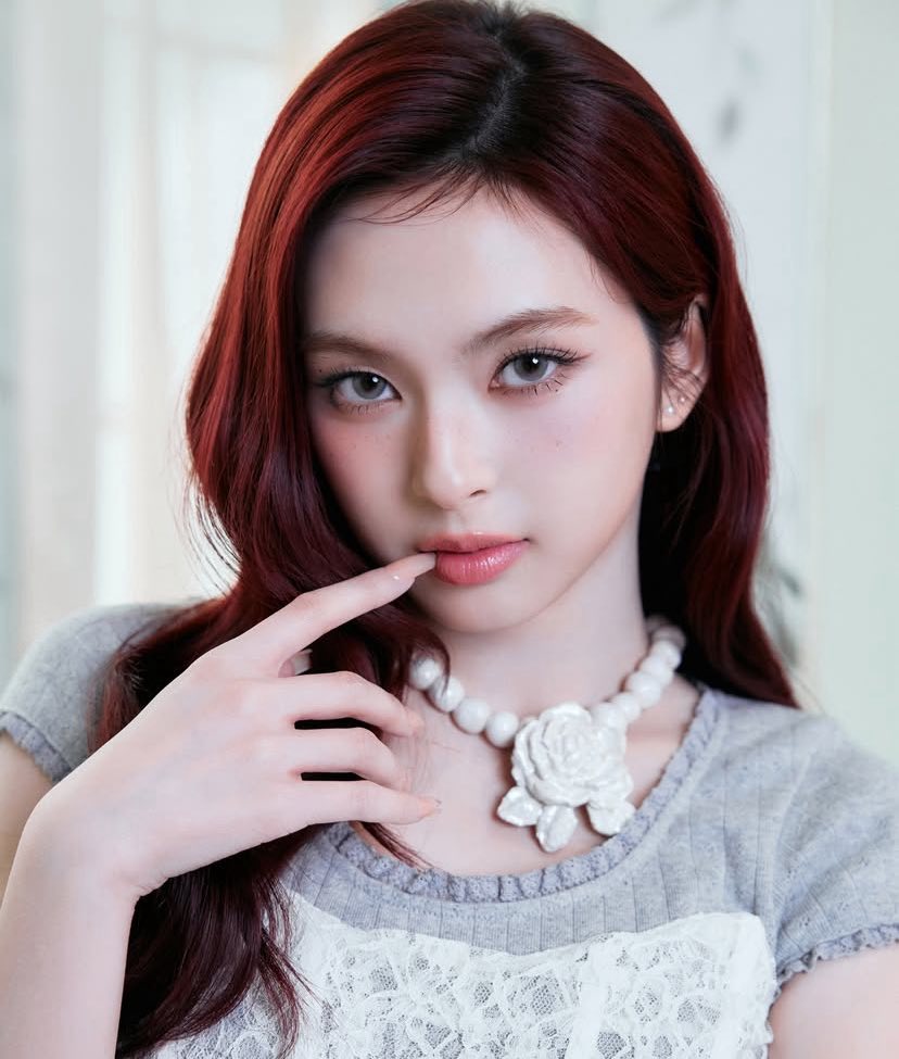 Young woman with dark red hair wearing white lace top and pearl flower necklace touching her lip