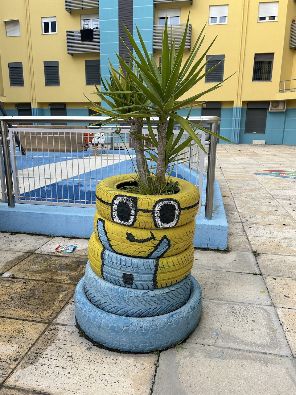 Stacked tires painted as a Minion character holding a palm plant in a colorful urban courtyard