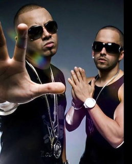 Two men in sunglasses and jewelry posing together against a dark background
