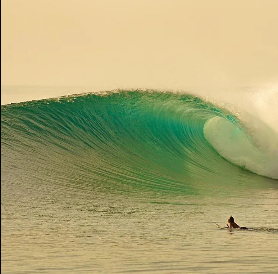 Large turquoise wave curling with surfer in calm ocean water under warm sky