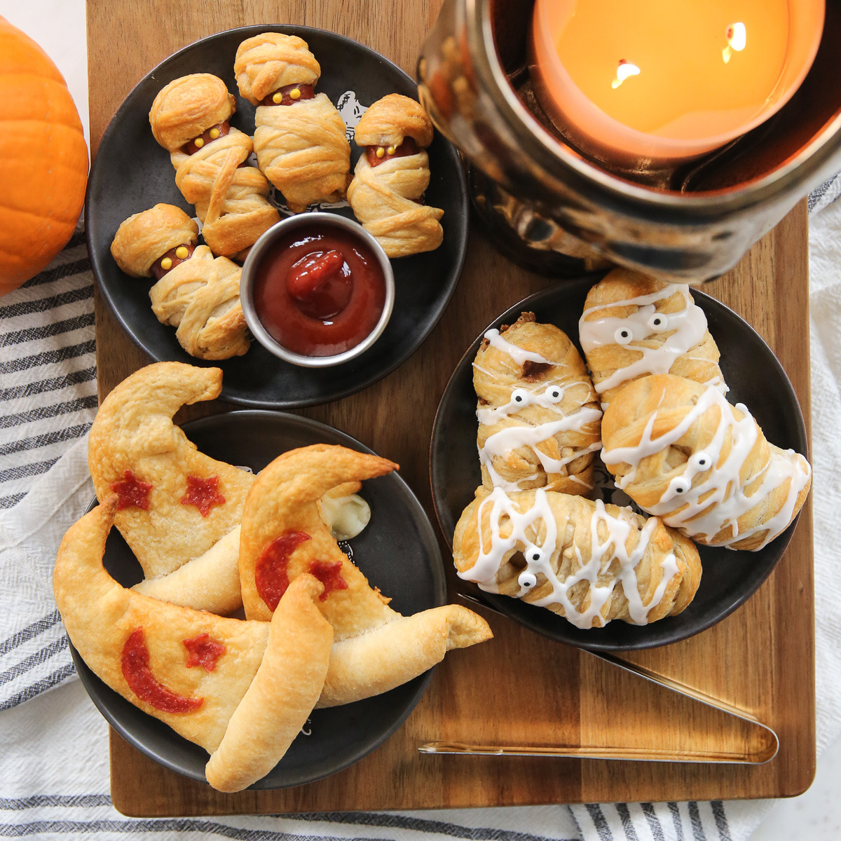 Halloween themed party food with mummy wrapped hot dogs, pastries shaped like pumpkins, and dipping sauces on black plates