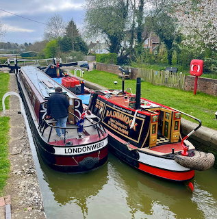 Two narrowboats moored on a canal beside a grassy towpath with trees and cloudy sky