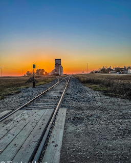 Railroad tracks converging toward a grain elevator at sunset on flat prairie landscape