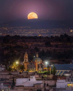 Large golden moon rising over a Mexican town with an illuminated church and distant city lights at night