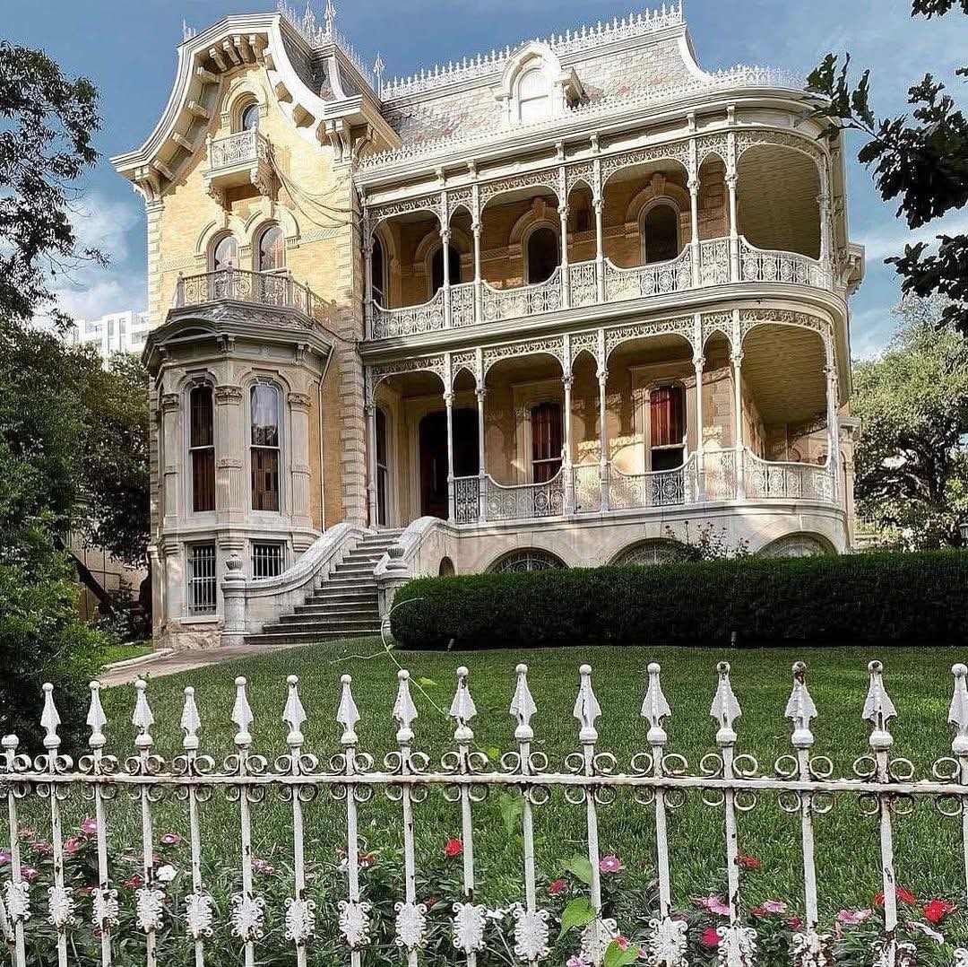Ornate Victorian house behind a white iron fence with hedges and front steps