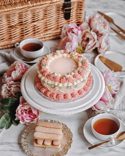 Pink decorated layer cake with tea cups, pink flowers, and wafer cookies on white picnic setup