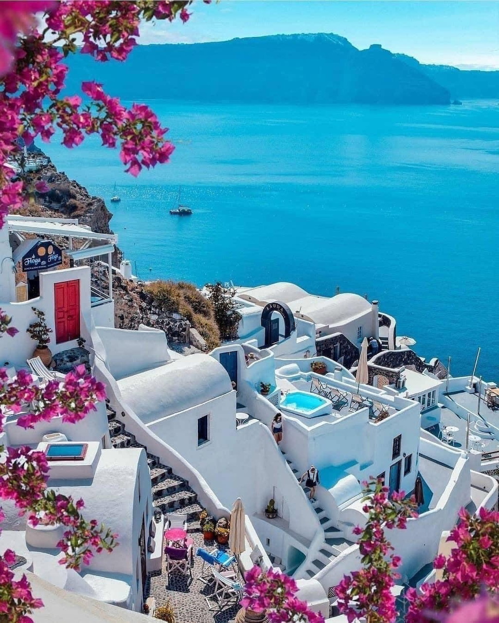 White-washed Santorini buildings on caldera cliffs framed by pink bougainvillea with blue Aegean Sea view