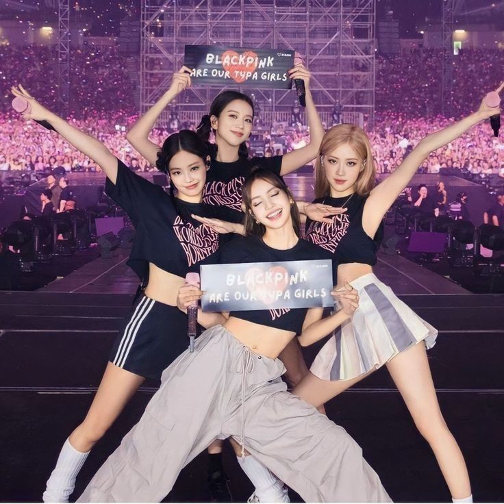 Four K-pop performers posing on stage with arms raised during a concert with purple stage lighting and crowd
