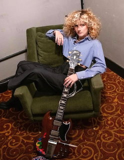 Young woman with curly blonde hair sitting in green armchair holding a black electric guitar
