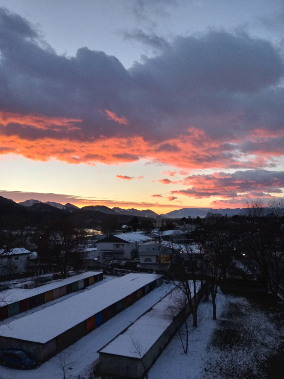 Winter sunset over a snowy village with rooftops, trees, and distant hills under dark clouds