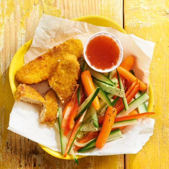 Plate of golden chicken strips with fresh vegetable sticks and red dipping sauce on yellow wooden table