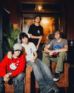 Four young band members posing indoors, one holding a red electric guitar, seated on leather furniture