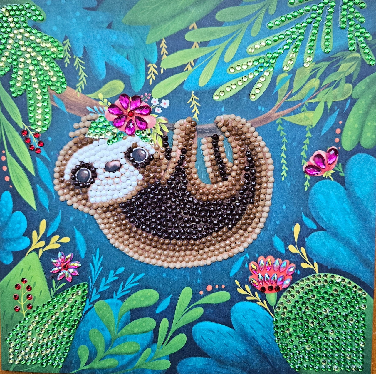 Beaded craft art showing a brown sloth with flowers hanging from green leaves on blue background