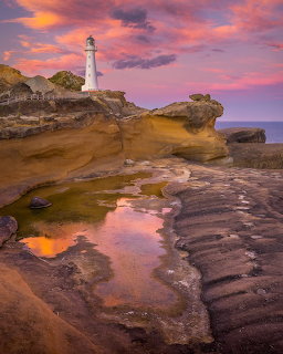 White lighthouse on rocky coast at sunset with pink clouds reflected in tidal pool among sandstone formations