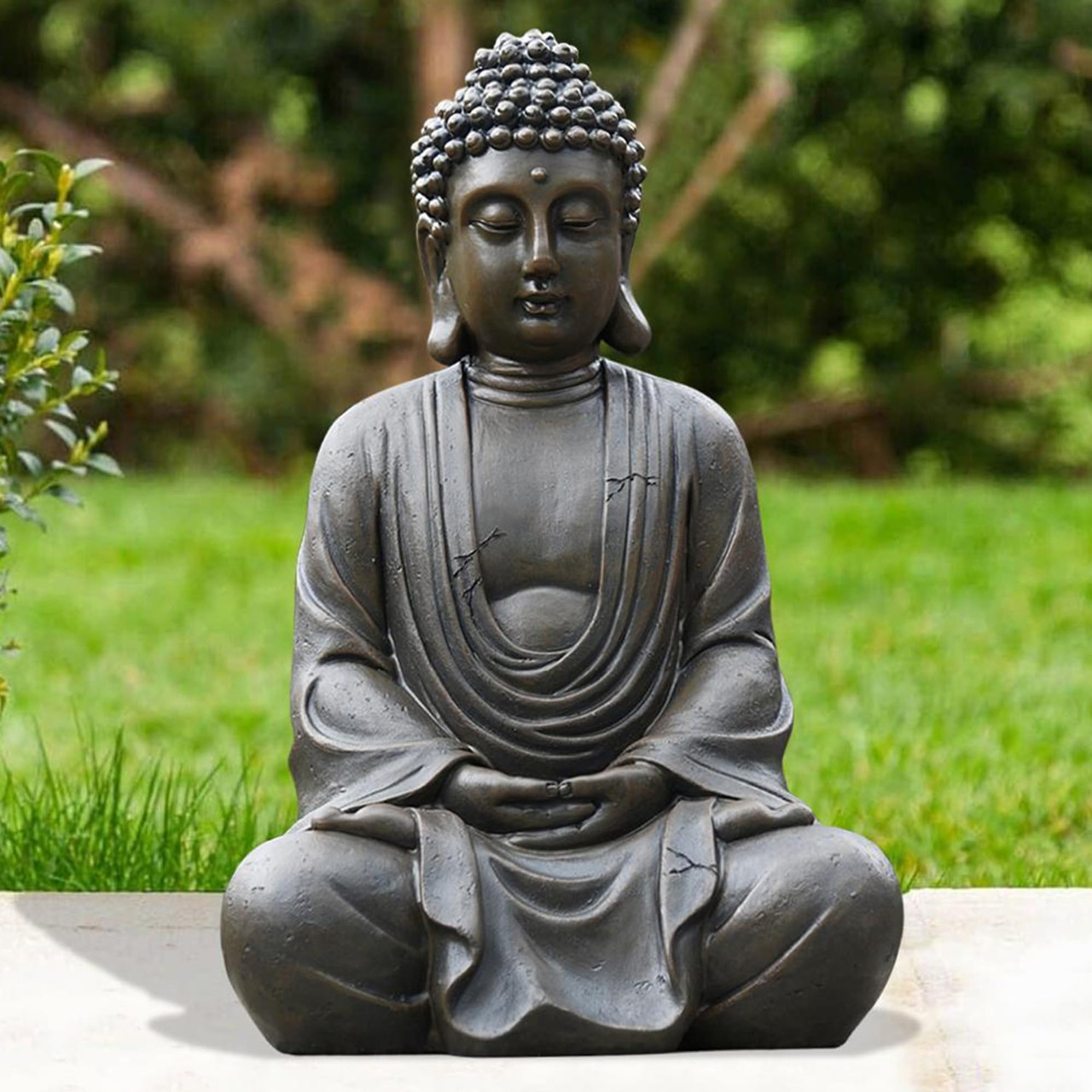 Gray Buddha statue sitting in lotus position outdoors with green garden background
