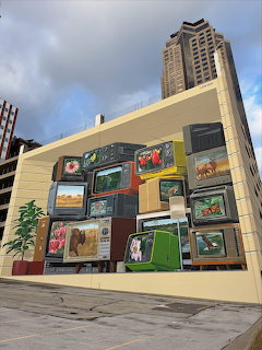Art installation of stacked vintage TV screens on a building wall with a tall skyscraper behind it