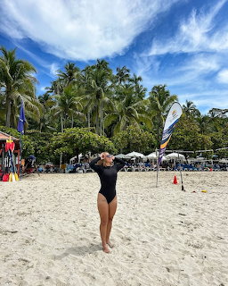 Woman in black swimsuit standing on beach holding white surfboard under palm trees and blue sky