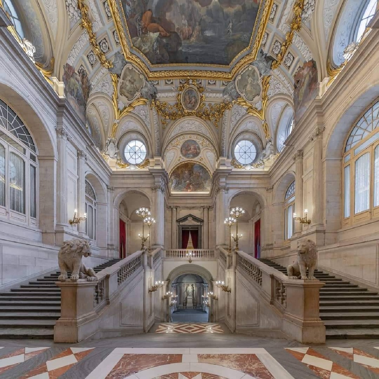 Grand ornate hall with twin marble staircases, statues, tall windows, and a painted ceiling