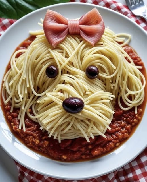 Spaghetti arranged on a plate to resemble a dog face with olive eyes and a ham bow on a checkered tablecloth