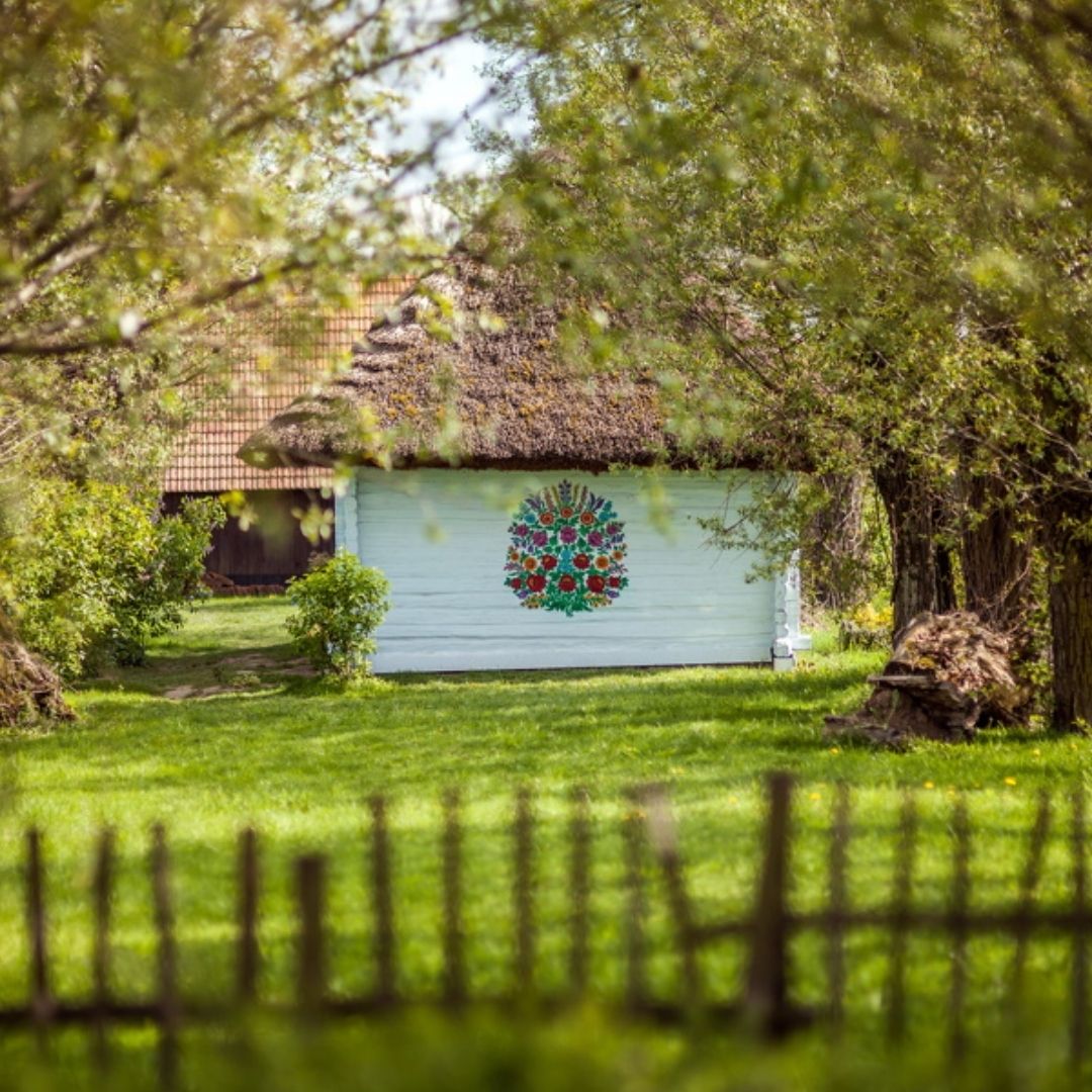White cottage with tiled roof behind a wooden fence, surrounded by trees and green lawn