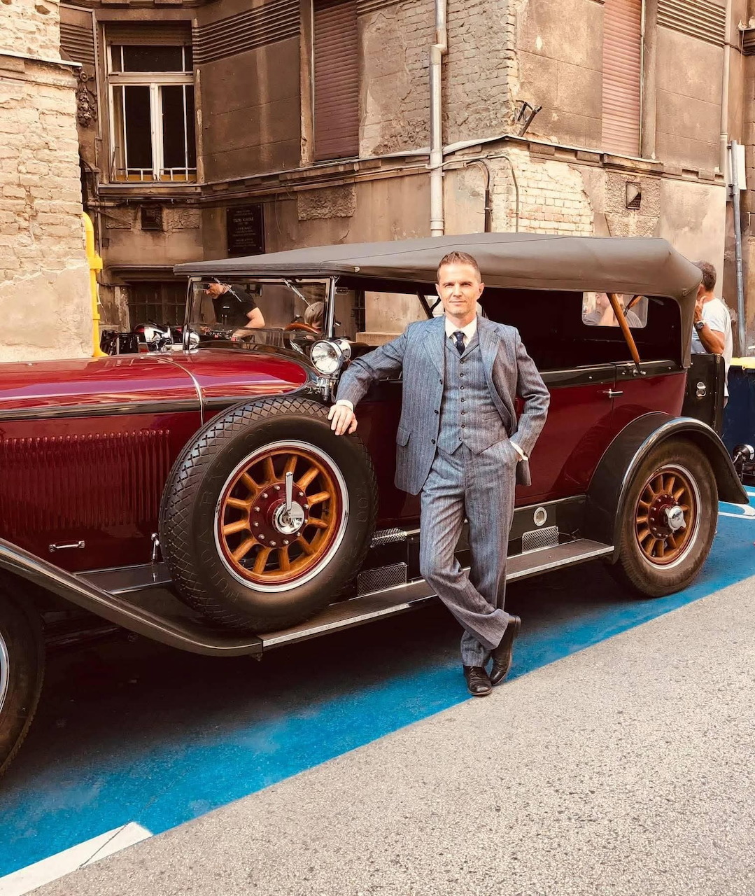 Man in grey suit posing next to a burgundy vintage car on a city street