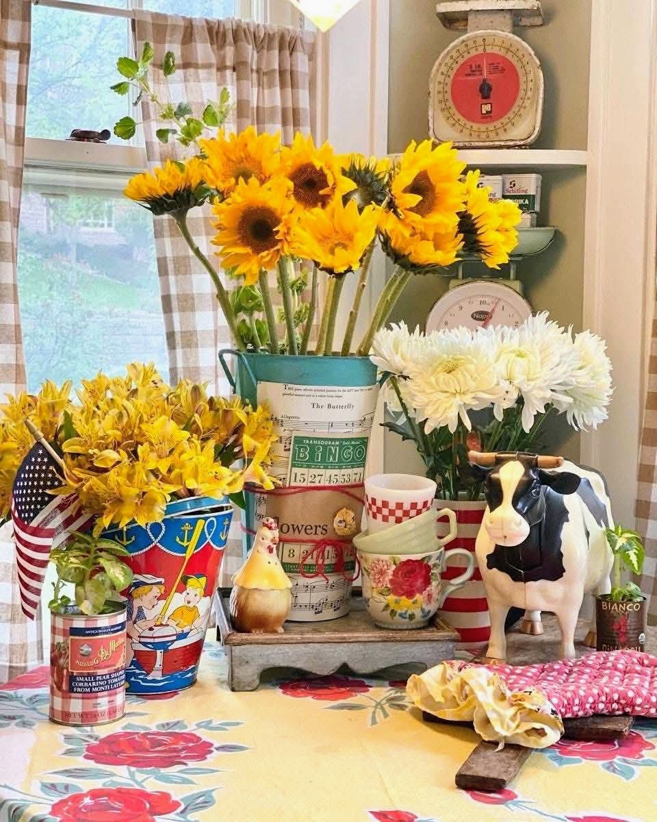 Vintage kitchen display with sunflowers, white flowers, mugs, a cow pitcher, and a red scale near a window