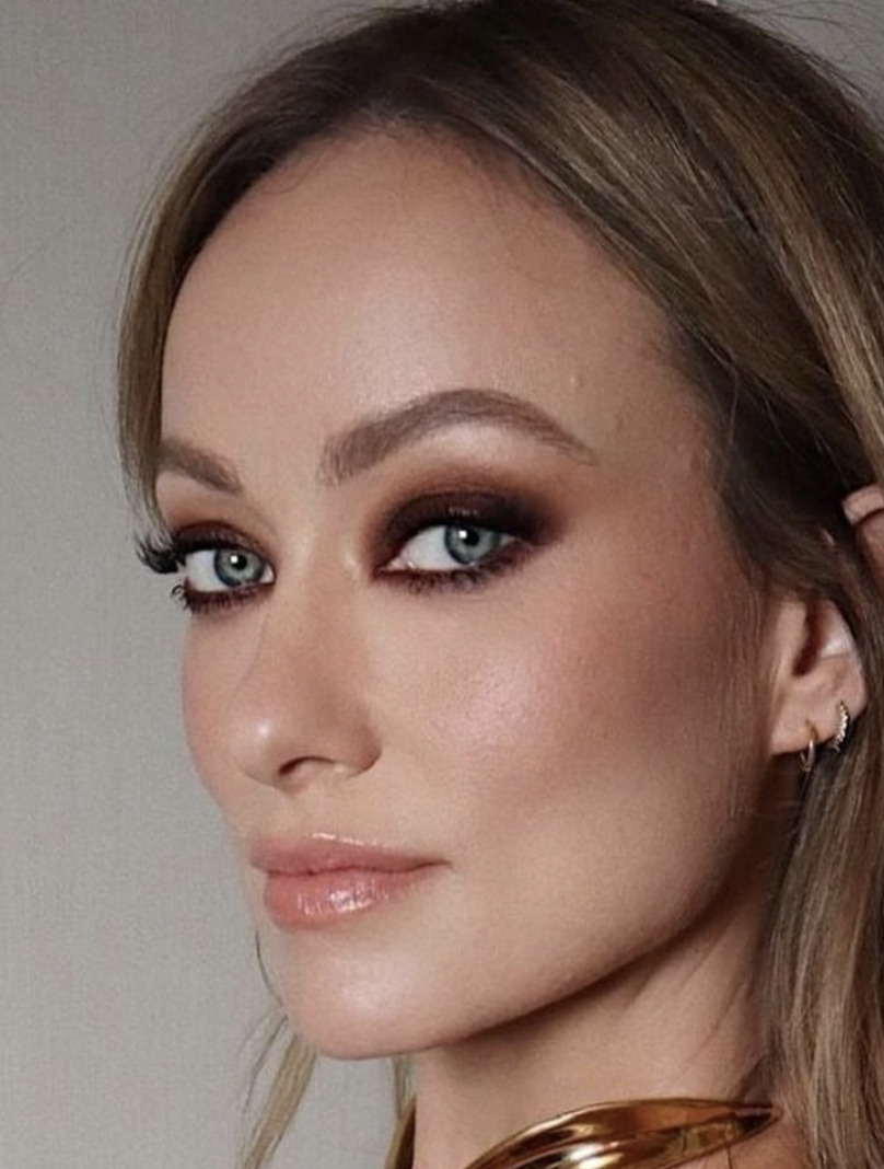 Close-up of a woman with smoky eye makeup, blue eyes, and gold jewelry against a neutral background.