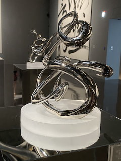 Abstract black steel sculpture with curved flowing forms on white pedestal in gallery