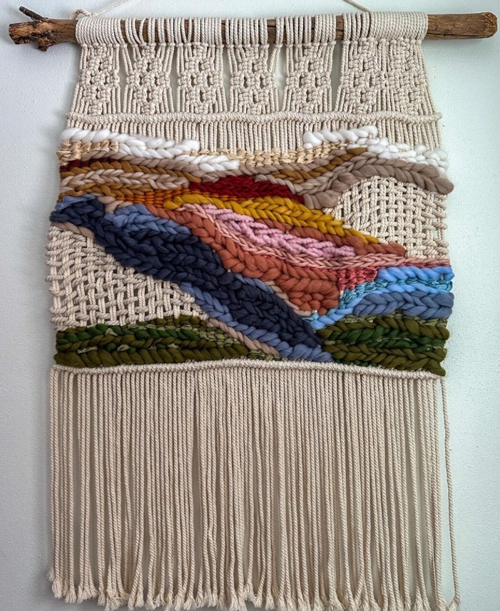Macrame wall hanging with colorful woven yarn sections in pink, blue, mustard, and green, suspended from a wooden stick.
