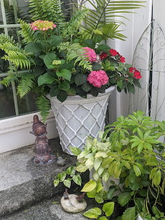 White geometric planter with pink flowers and green plants on a porch surrounded by ferns and foliage
