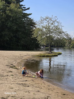 Two young children playing in the sand on a riverbank with trees and water behind them