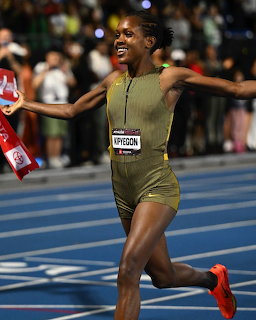 Female runner in gold uniform celebrates crossing finish line with arms spread wide on blue track