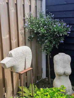 Two white abstract stone sculptures in a garden with plants and wooden fence