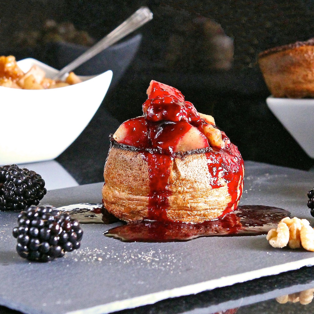 Golden pastry drizzled with red berry compote, surrounded by blackberries and hazelnuts on slate