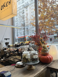 Cafe counter by a window with jars, pastries, pumpkins, and autumn decorations overlooking a city street.