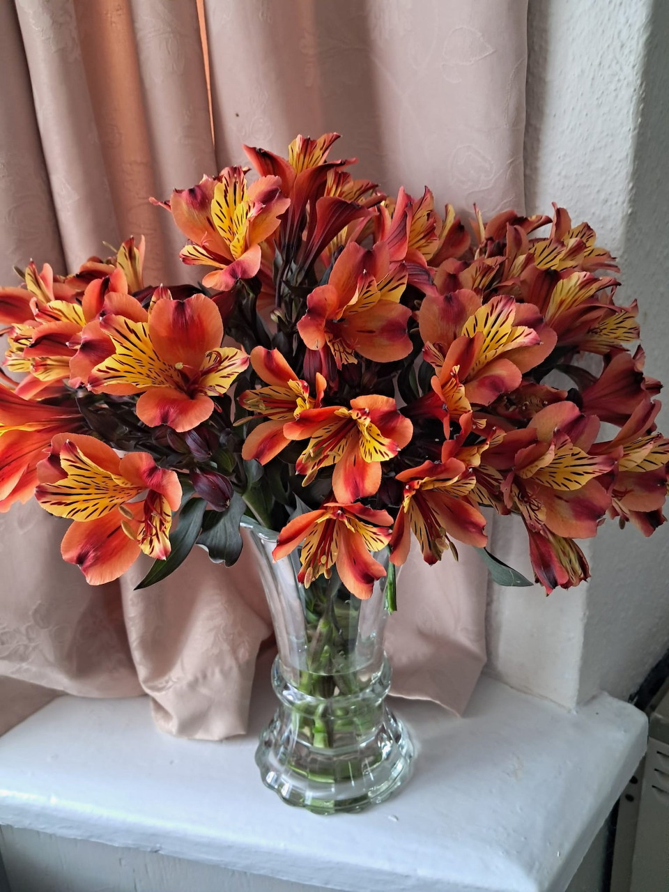 Glass vase of orange and yellow alstroemeria flowers on a white surface by a pink curtain