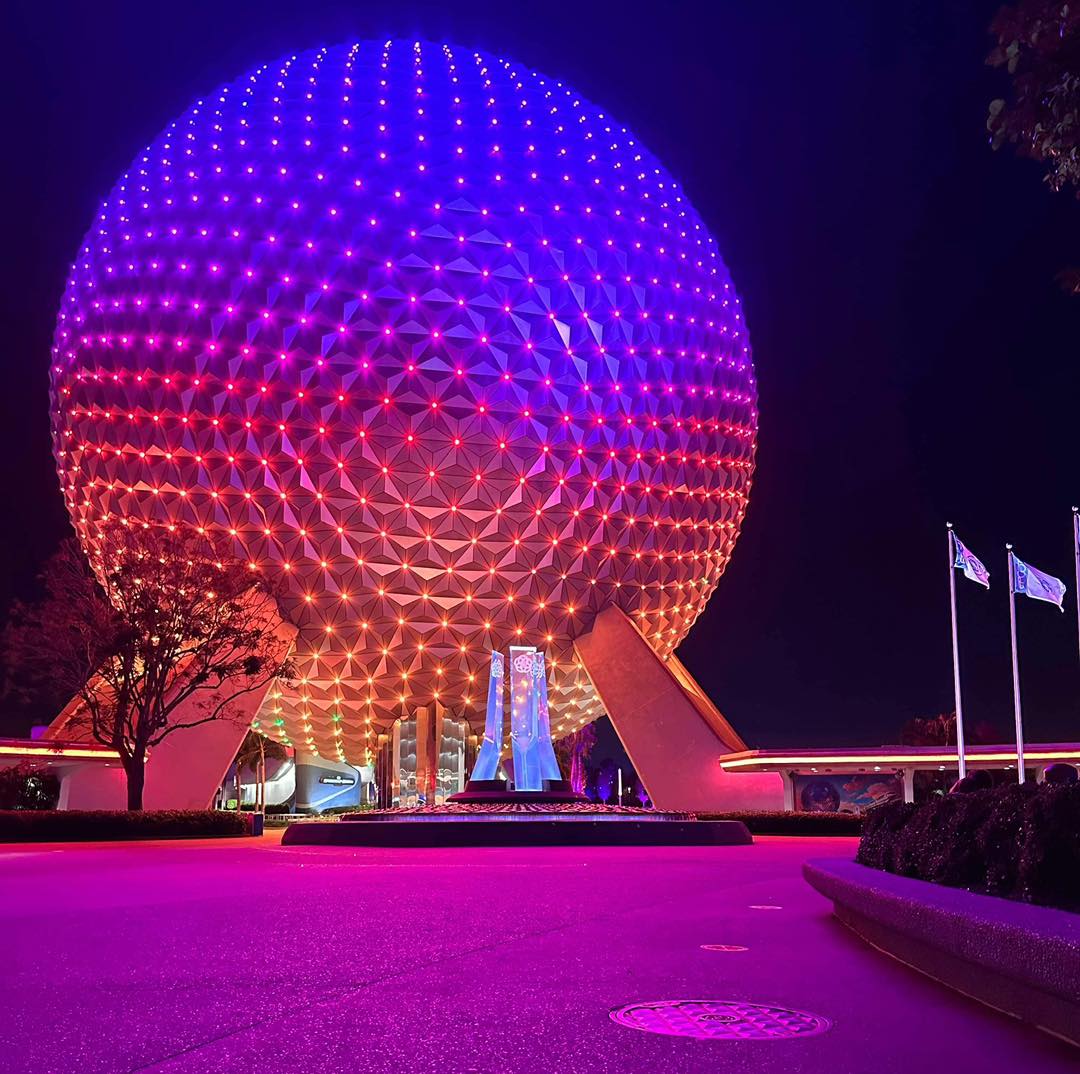 Spaceship Earth lit in purple and orange at night with flags and a walkway in front