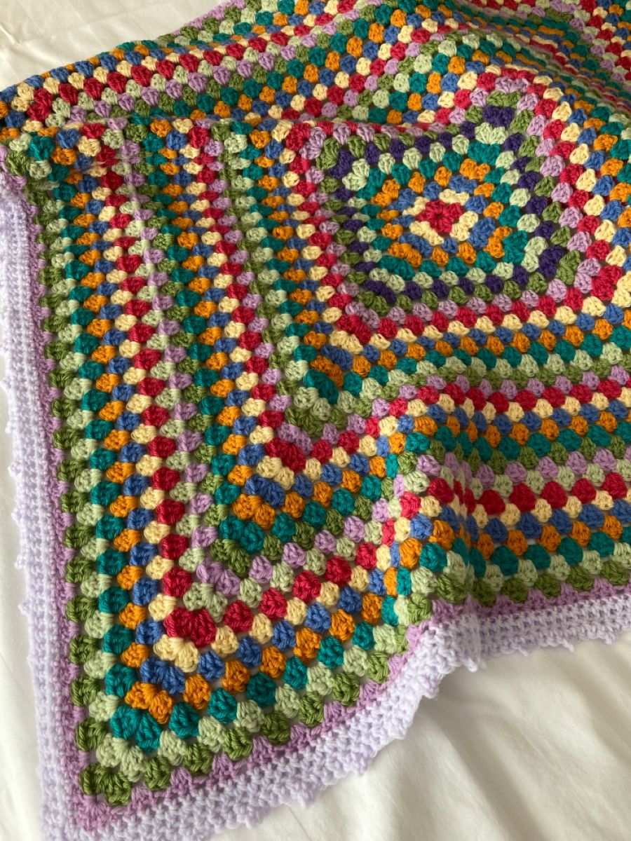 Multicolored crochet granny square blanket with lavender border laid flat