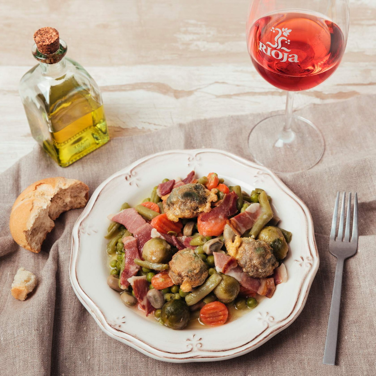 Plate of gnocchi with vegetables, olives, bread, olive oil bottle, and glass of red wine on beige linen tablecloth