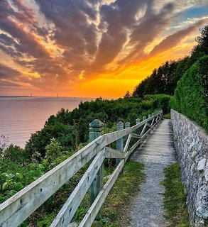 Wooden walkway along a stone wall with green trees overlooking water at a vibrant orange sunset